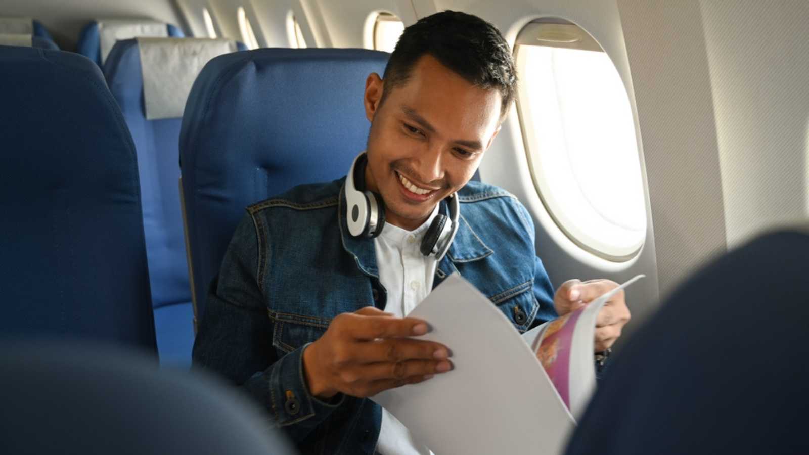 Cheerful asian man passenger in hipster outfits sitting near window in an airplane and reading magazine