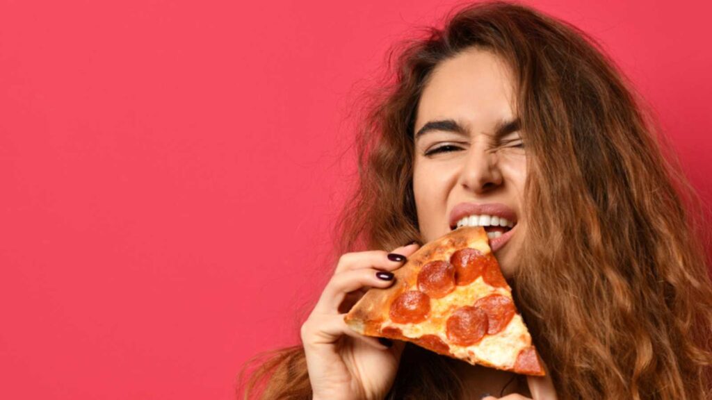 Young beautiful woman with slice of pepperoni pizza screaming laughing on dark pink background