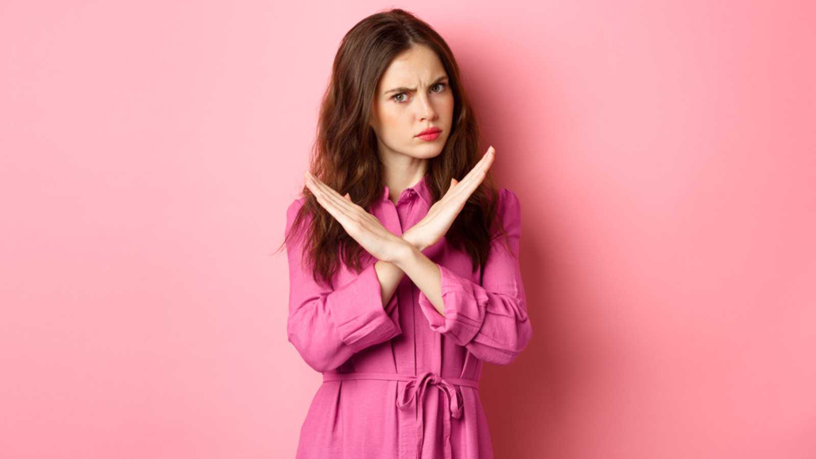 Displeased angry woman blocking offer, showing cross stop gesture, saying no and shaking head in negative reply, standing over pink background