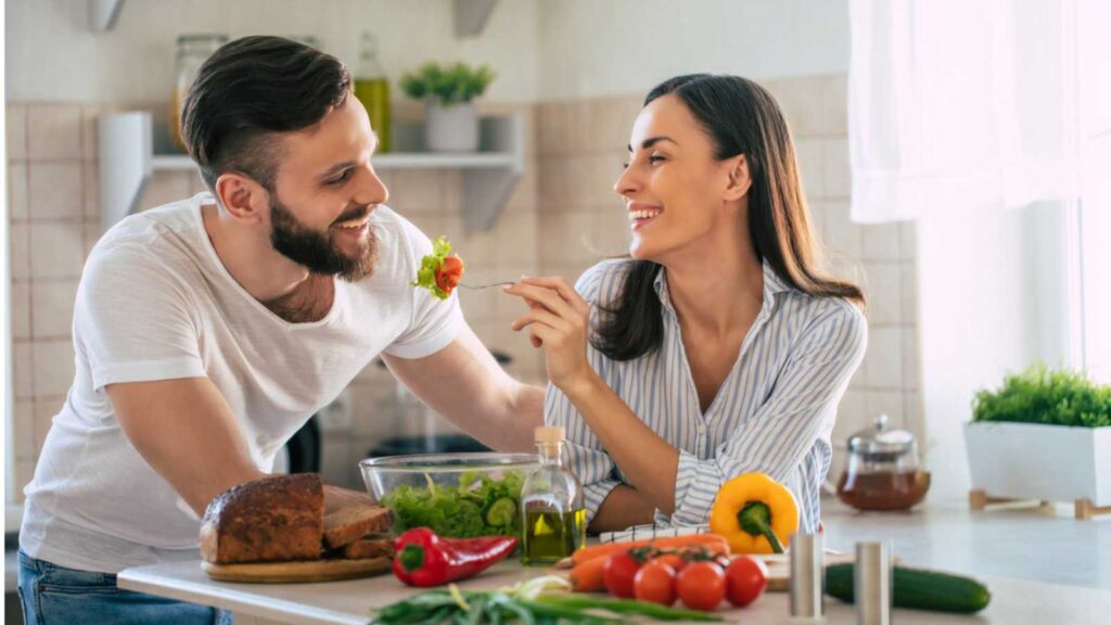 Excited smiling young couple in love making a super healthy vegan salad with many vegetables in the kitchen and man testing it from a girl's hands