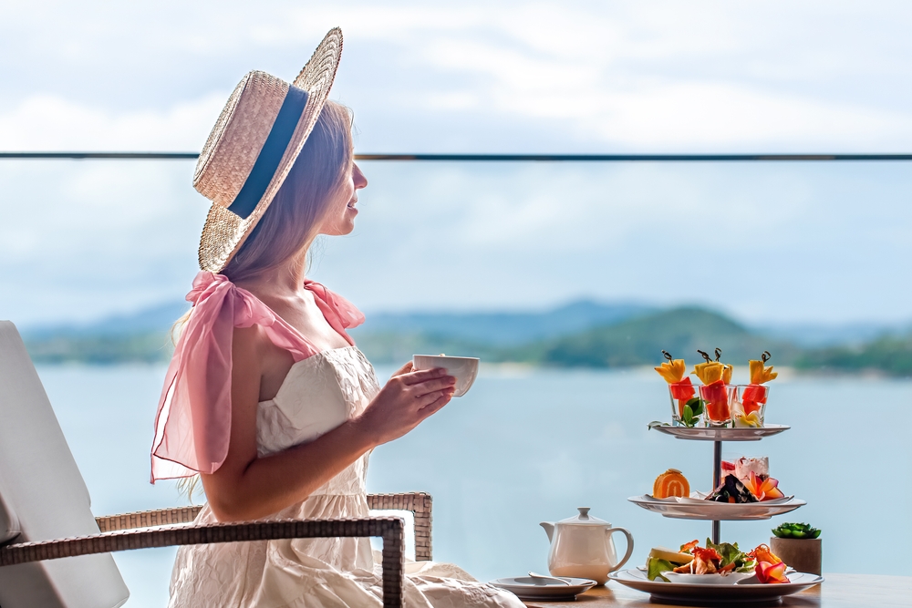 Young lady in white dress drinking tea on Afternoon tea with mini canape and selection of various sweets with sea on background.