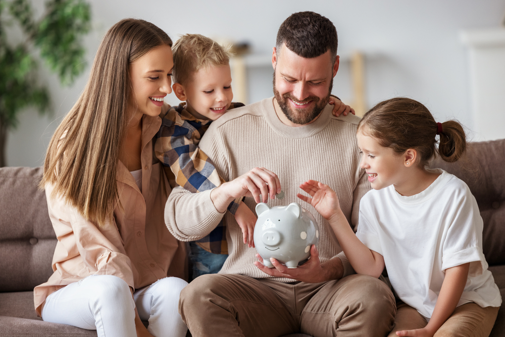 Happy family: cheerful mother and father with kids smiling and putting coins into piggy bank while sitting on sofa at home.